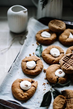 A tray lined with parchment paper holds freshly baked cookies. Each cookie features a distinct pattern and is topped with a small, white marshmallow. Nearby, an empty, white ceramic jug adds a touch of simplicity to the scene. Crumbs are lightly scattered around, emphasizing the fresh, homemade feel. A couple of green leaves are placed on the tray, adding a natural element to the display.