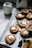 A tray lined with parchment paper holds freshly baked cookies. Each cookie features a distinct pattern and is topped with a small, white marshmallow. Nearby, an empty, white ceramic jug adds a touch of simplicity to the scene. Crumbs are lightly scattered around, emphasizing the fresh, homemade feel. A couple of green leaves are placed on the tray, adding a natural element to the display.