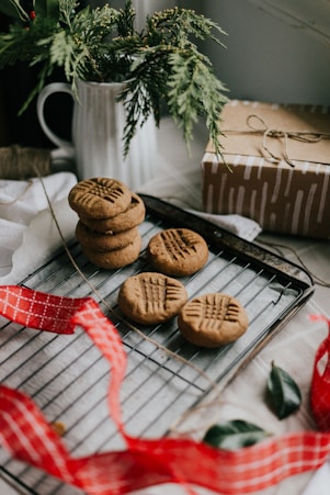 A set of freshly baked cookies with a pattern on top is placed on a cooling rack. The scene includes a red checkered ribbon, some greenery in a white ceramic container, and a brown gift box wrapped with twine, contributing to a festive and cozy atmosphere.