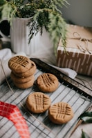 A wooden table displays a baking rack with several peanut butter cookies. The cookies have a distinct criss-cross pattern on top and are fresh out of the oven. A white pitcher with evergreen branches adds a rustic touch, while a wrapped gift box tied with twine sits nearby. A red ribbon is partially visible, adding a hint of color to the scene.