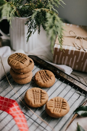 A wooden table displays a baking rack with several peanut butter cookies. The cookies have a distinct criss-cross pattern on top and are fresh out of the oven. A white pitcher with evergreen branches adds a rustic touch, while a wrapped gift box tied with twine sits nearby. A red ribbon is partially visible, adding a hint of color to the scene.