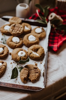 A baking sheet with eight cookies, some topped with a marshmallow or a similar white topping, arranged on parchment paper. The cookies appear to be freshly baked, with one cookie partly bitten. There are green leaves placed decoratively on the baking sheet and a red-and-black checkered cloth in the background.