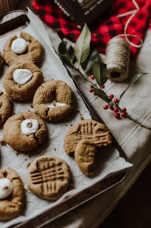 A baking tray filled with freshly baked cookies featuring various textures and designs, including some topped with marshmallows. Surrounding the tray are holiday-themed decorations such as branches with red berries, a roll of twine, and a red checkered cloth, creating a cozy and festive atmosphere.