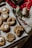 A baking tray filled with freshly baked cookies featuring various textures and designs, including some topped with marshmallows. Surrounding the tray are holiday-themed decorations such as branches with red berries, a roll of twine, and a red checkered cloth, creating a cozy and festive atmosphere.