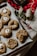 A baking tray filled with freshly baked cookies featuring various textures and designs, including some topped with marshmallows. Surrounding the tray are holiday-themed decorations such as branches with red berries, a roll of twine, and a red checkered cloth, creating a cozy and festive atmosphere.