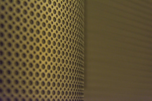 A close-up view of a perforated metal surface with a pattern of evenly spaced circular holes. The lighting casts a shadow that adds depth and creates a gradient effect across the surface.