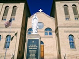 A historical church with a beige brick facade features a statue of a religious figure in white, standing on a plinth inscribed with names. Stained glass windows with intricate patterns are located above the entrance. An American flag is visible to the side.