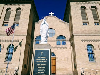 A historical church with a beige brick facade features a statue of a religious figure in white, standing on a plinth inscribed with names. Stained glass windows with intricate patterns are located above the entrance. An American flag is visible to the side.