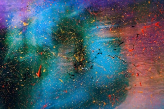 An abstract artwork with swirling colors and dynamic splatters. Vivid blue and green tones blend into shades of purple and red, with scattered dots and streaks of yellow, orange, and black adding texture and movement.