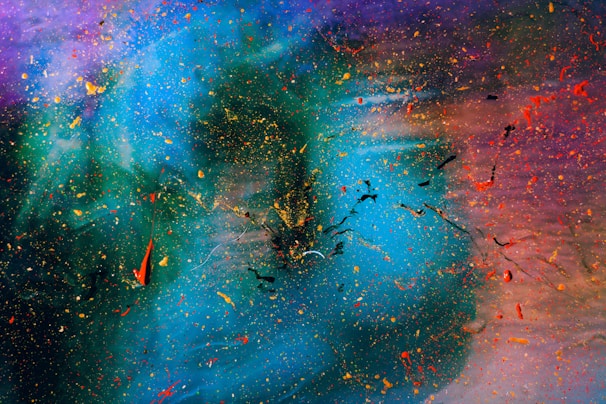 An abstract artwork with swirling colors and dynamic splatters. Vivid blue and green tones blend into shades of purple and red, with scattered dots and streaks of yellow, orange, and black adding texture and movement.