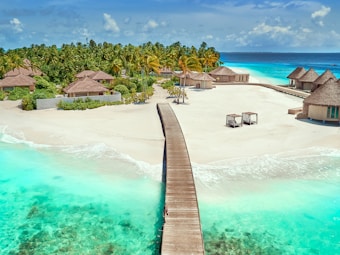 A scenic tropical beach scene with turquoise waters and a long wooden boardwalk leading to a cluster of thatched-roof huts. Lush green palm trees line the top edge of the sand, and white sandy beaches stretch under a clear blue sky.