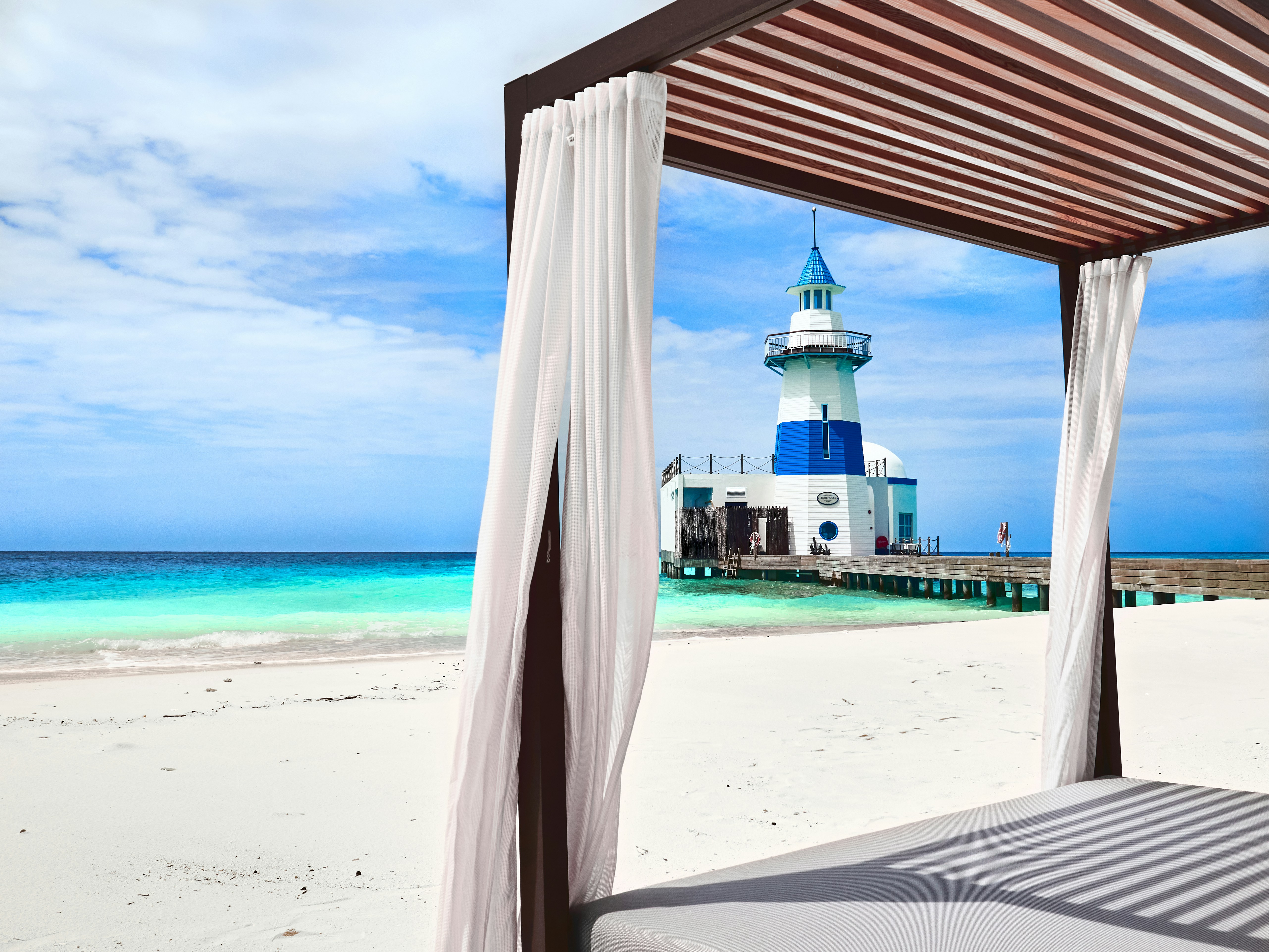 Luxury beach cabana with white curtains, comfortable daybed, tropical palm trees, dappled light