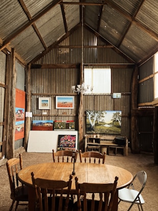 A rustic wooden barn interior with corrugated metal walls, featuring an assortment of paintings and artwork displayed on the walls and floor. A wooden round table with wooden chairs is positioned in the center of the room. The artworks include landscapes and abstract paintings. A chandelier hangs from the ceiling, adding a touch of elegance to the rustic setting.