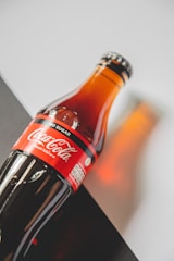 A glass bottle of Coca-Cola with a red label marked 'No Sugar'. The bottle is placed on a diagonal, set against a background with contrasting light and dark areas. The lighting creates a warm reflection on the surface.