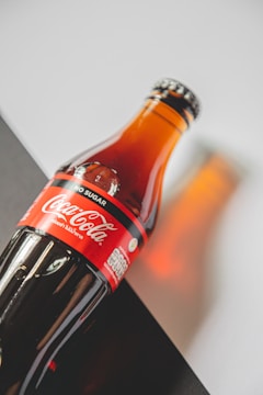 A glass bottle of Coca-Cola with a red label marked 'No Sugar'. The bottle is placed on a diagonal, set against a background with contrasting light and dark areas. The lighting creates a warm reflection on the surface.