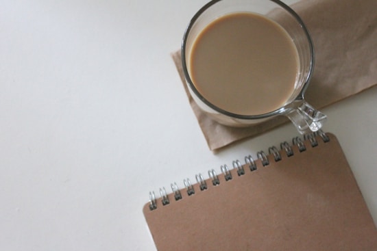 A glass mug filled with coffee or tea sits atop a brown paper napkin on a white surface. Next to the mug is a spiral-bound, kraft paper-covered notebook.