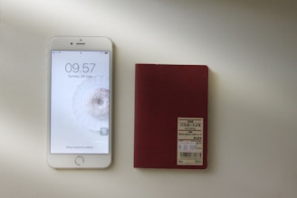 A smartphone with a visible lock screen displaying the time, date, and a motivational background is placed next to a red notepad with a label containing text in Japanese. Both items are situated on a light surface with a shadow cast across them.