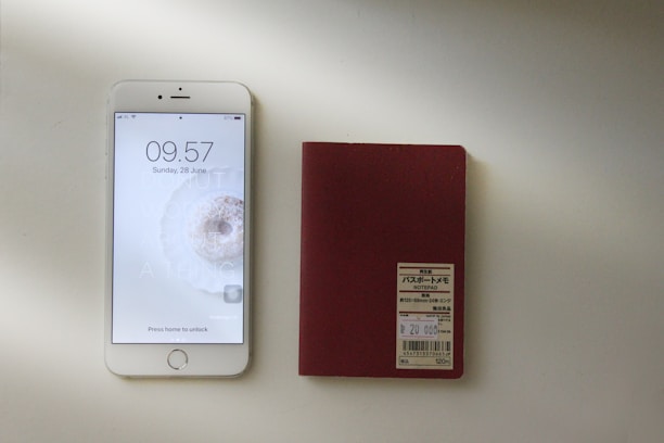 A smartphone with a visible lock screen displaying the time, date, and a motivational background is placed next to a red notepad with a label containing text in Japanese. Both items are situated on a light surface with a shadow cast across them.