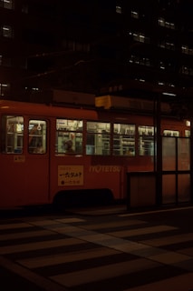 A dimly-lit streetcar is stationary at a stop in an urban environment. The streetcar features a vibrant red-orange exterior with illuminated windows showing passengers inside. The background consists of a dark building with numerous small lit windows, while the foreground includes a striped pedestrian crossing.
