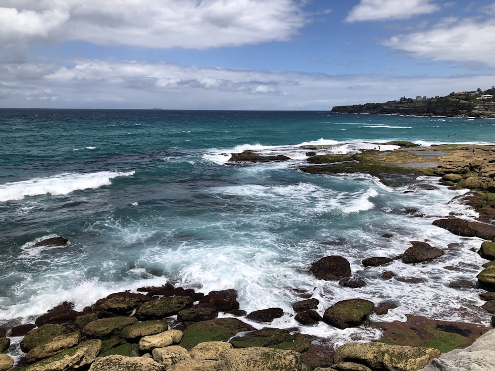 olas oceánicas rompiendo contra las rocas bajo el cielo nublado azul y blanco durante el día
