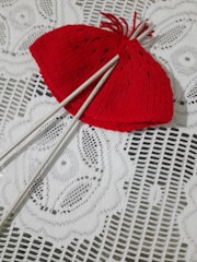 A vibrant red knitted hat is placed on a white lace tablecloth. Two white knitting needles are inserted through the hat. The lace tablecloth has an intricate floral pattern.