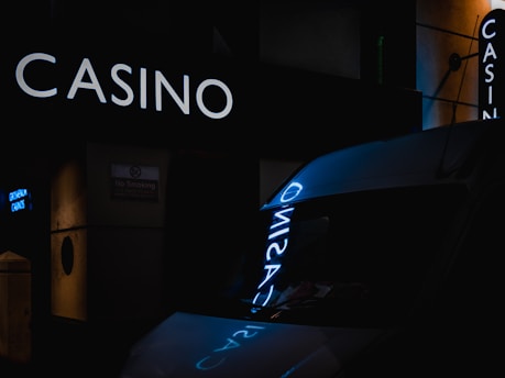 A dimly lit urban scene with a prominent glowing casino sign reflecting off a nearby surface. The setting appears to be at night, with dark shadows and minimal lighting elsewhere. A 'No Smoking' sign is partially visible on one side.