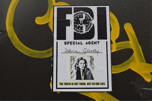 A poster-like image of an FBI special agent identification card with the name Dana Scully. The card features an official seal and a black-and-white photo of a woman with shoulder-length hair. Below the photo is a quote: 'The truth is out there, but so are lies.' The background includes yellow graffiti-like markings.