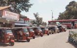 Several tuk-tuks are parked in a row alongside a street near a small shop labeled 'Highland Chathurani Milk Bar' with a red and white striped sign. A red bus is visible in the background, with a few people walking nearby. The setting appears to be in a bustling, sunny area with some trees and greenery around.