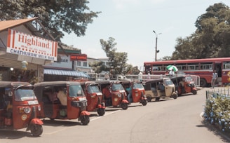 Several tuk-tuks are parked in a row alongside a street near a small shop labeled 'Highland Chathurani Milk Bar' with a red and white striped sign. A red bus is visible in the background, with a few people walking nearby. The setting appears to be in a bustling, sunny area with some trees and greenery around.