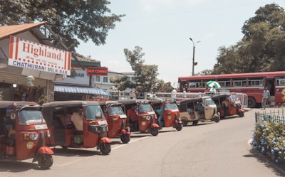 Several tuk-tuks are parked in a row alongside a street near a small shop labeled 'Highland Chathurani Milk Bar' with a red and white striped sign. A red bus is visible in the background, with a few people walking nearby. The setting appears to be in a bustling, sunny area with some trees and greenery around.
