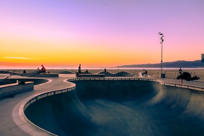 A skate park near the beach at sunset, with people skating on the smooth concrete surfaces. The sky is colorful, transitioning from pink and purple to golden orange as the sun sets over the horizon. An ocean and distant mountains are visible in the background, providing a scenic backdrop. The park is lit by tall lights, and some skaters are performing tricks while others are relaxing.