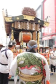 A decorated float is pulled through a festival with detailed wooden carvings and traditional Japanese elements. Several people in traditional attire, including a person wearing a garment with a dragon pattern, accompany the float. Banners with Japanese writing hang from the float, and a large drum is visible in the center.