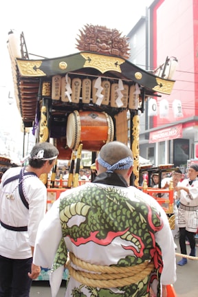A decorated float is pulled through a festival with detailed wooden carvings and traditional Japanese elements. Several people in traditional attire, including a person wearing a garment with a dragon pattern, accompany the float. Banners with Japanese writing hang from the float, and a large drum is visible in the center.