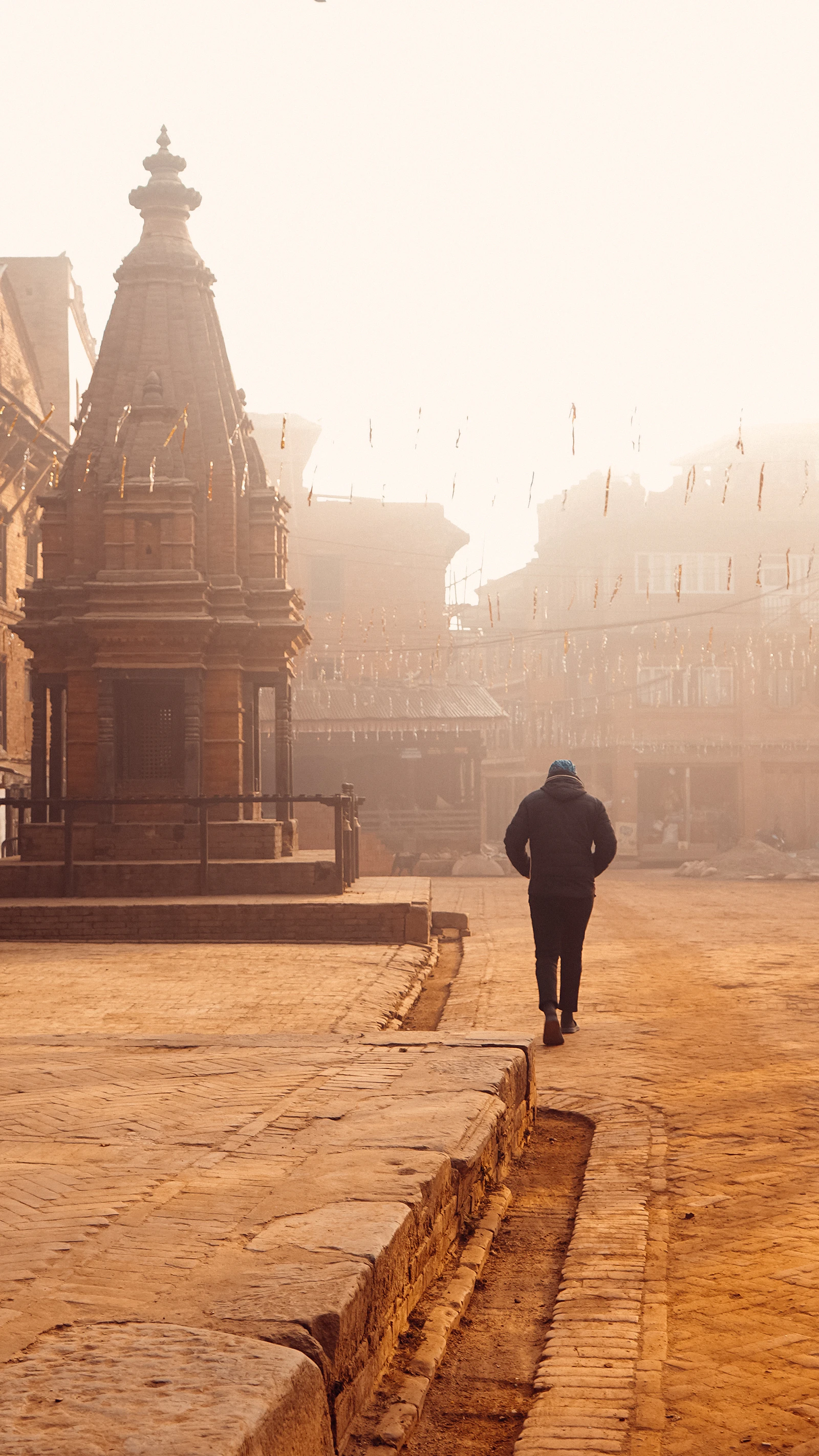 Morning fog in Panauti's old square, a brick shikhara temple on the left and a lone walker heading toward the prayer flags