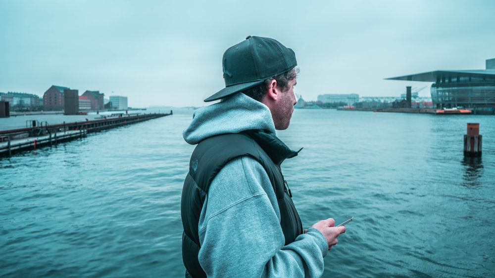man in gray hoodie and green cap standing near body of water during daytime