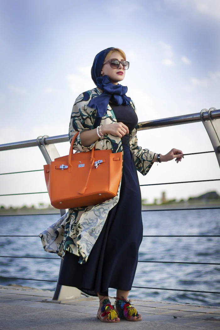 Femme élégante en hijab de soie bleu nuit, manteau imprimé sur robe marine et sac orange, au bord de l'eau