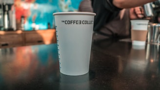 A disposable coffee cup with 'THE COFFEE COLLECTIVE' printed on it sits on a table. In the background, there's a person preparing a drink and another takeaway cup. The setting appears to be a coffee shop.