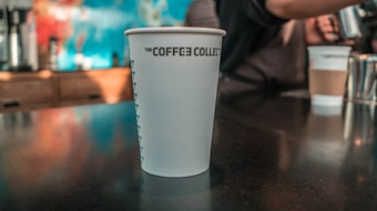 A disposable coffee cup with 'THE COFFEE COLLECTIVE' printed on it sits on a table. In the background, there's a person preparing a drink and another takeaway cup. The setting appears to be a coffee shop.
