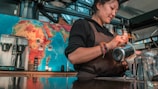 A barista wearing a brown apron is skillfully pouring milk into a coffee cup. Behind her, a colorful world map adorns the wall, and various coffee-making equipment is arranged on the counter.
