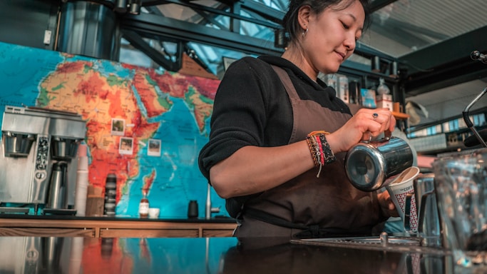 A barista wearing a brown apron is skillfully pouring milk into a coffee cup. Behind her, a colorful world map adorns the wall, and various coffee-making equipment is arranged on the counter.