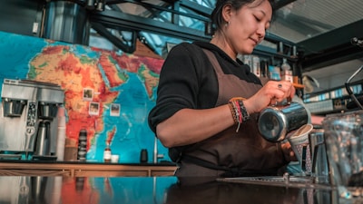 A barista wearing a brown apron is skillfully pouring milk into a coffee cup. Behind her, a colorful world map adorns the wall, and various coffee-making equipment is arranged on the counter.