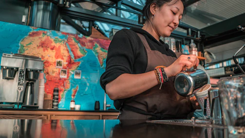A barista wearing a brown apron is skillfully pouring milk into a coffee cup. Behind her, a colorful world map adorns the wall, and various coffee-making equipment is arranged on the counter.