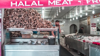 A butcher shop displays a large pile of animal skulls prominently at the front. There is a man talking on the phone inside, standing next to several trays of meat and fish arranged on counters. The shop has bright overhead lighting with numerous bulbs and a sign reads 'HALAL MEAT' in bold letters above the entrance.
