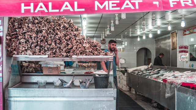 A butcher shop displays a large pile of animal skulls prominently at the front. There is a man talking on the phone inside, standing next to several trays of meat and fish arranged on counters. The shop has bright overhead lighting with numerous bulbs and a sign reads 'HALAL MEAT' in bold letters above the entrance.