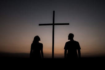 Silhouettes of a man and a woman are standing on either side of a large cross, with a clear evening sky in the background. The scene is serene and contemplative, with a soft gradient of colors transitioning from dark to light.
