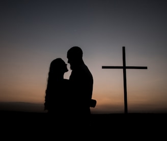 silhouette of man and woman standing beside cross during sunset