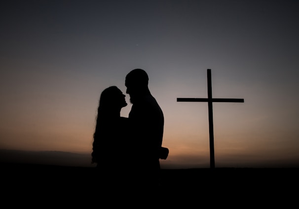 silhouette of man and woman standing beside cross during sunset