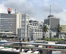 A historic-looking gray church with a clock tower stands prominently in the foreground, surrounded by modern high-rise buildings. Palm trees add a touch of greenery, and numerous market stalls or vehicles are seen below, suggesting a bustling urban environment.