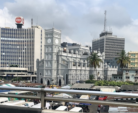 A historic-looking gray church with a clock tower stands prominently in the foreground, surrounded by modern high-rise buildings. Palm trees add a touch of greenery, and numerous market stalls or vehicles are seen below, suggesting a bustling urban environment.