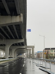 A wet road runs under a large, elevated highway or overpass structure. There are several vertical support columns holding up the overpass. Metal railings and road signs are visible along the road, which appears to be empty of vehicles. Accumulated rainwater is seen reflecting the surroundings. Residential buildings and streetlights are visible in the background.